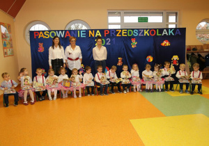 Grupa dzieci siedzi na krzesełkach, na kolanach trzymają upominki i dyplomy. Za dziećmi stoi pani dyrektor Maria Królikowska, pani Kinga i pani Ewelina.